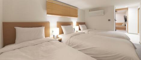 A hotel room with three beds, a wooden headboard, a window with blinds, a bedside table with a lamp, and a door leading to a bathroom.