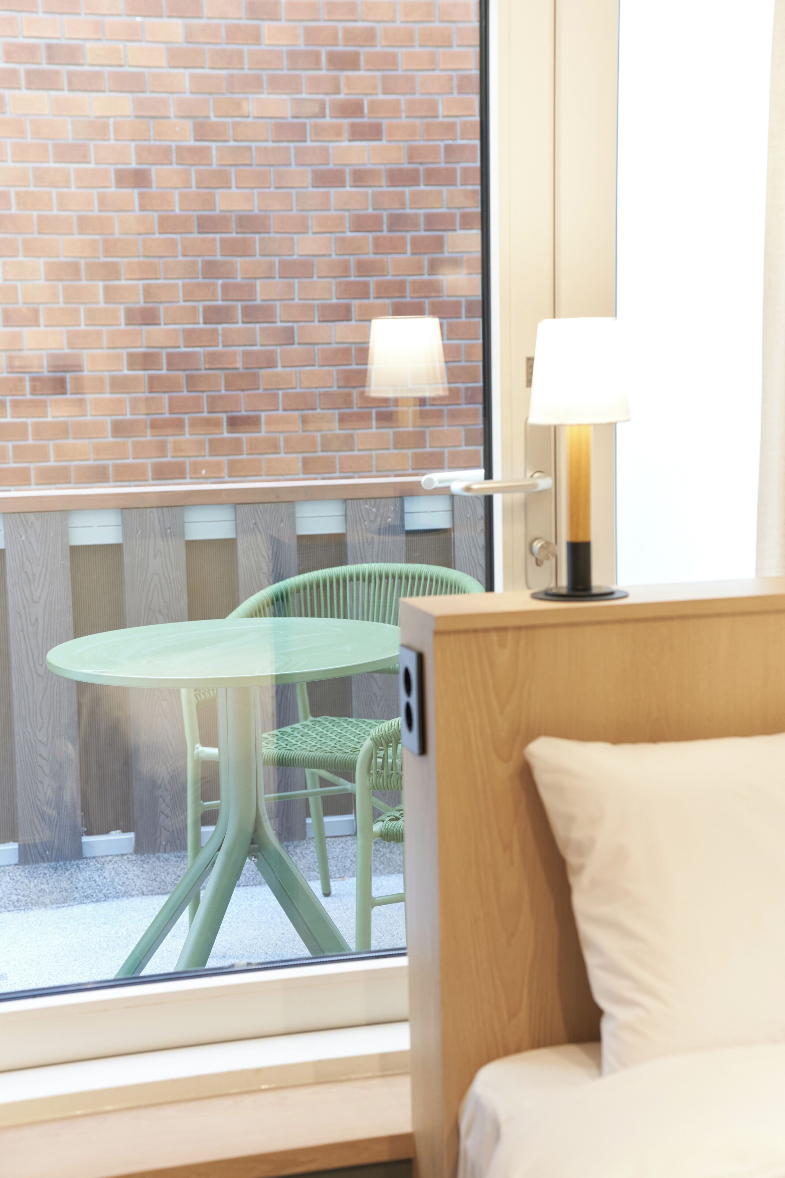 Een kamer met een houten hoofdbord, een groene stoel, een ronde tafel en uitzicht op een bakstenen muur door een raam.