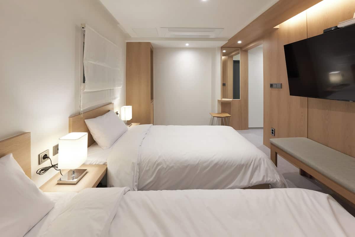 A hotel room with two beds, a TV, a bedside table with a lamp, and a bench.
