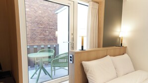 Habitación de hotel con cama, ventana que da a un edificio de ladrillo, pequeña mesa y una silla afuera.
