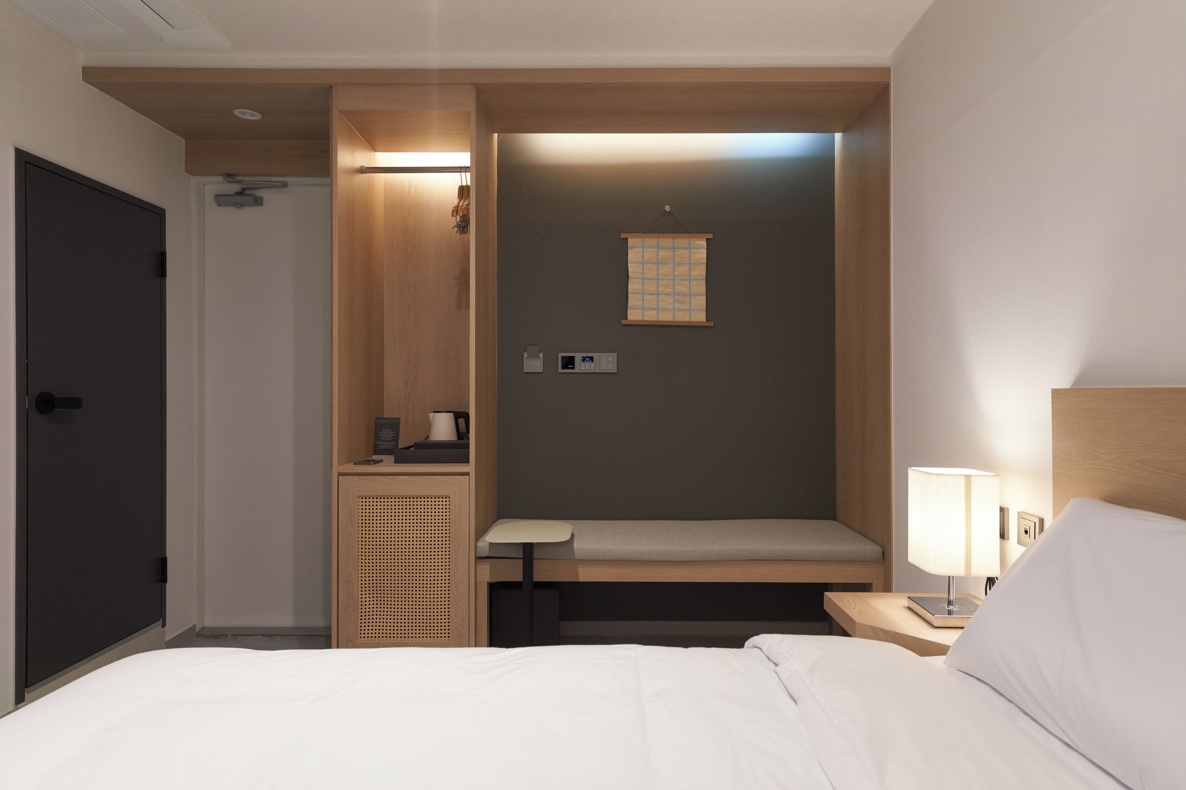 A hotel room with a bed, a bedside table with a lamp, a small bench, a wooden cabinet, and a wall-mounted panel.