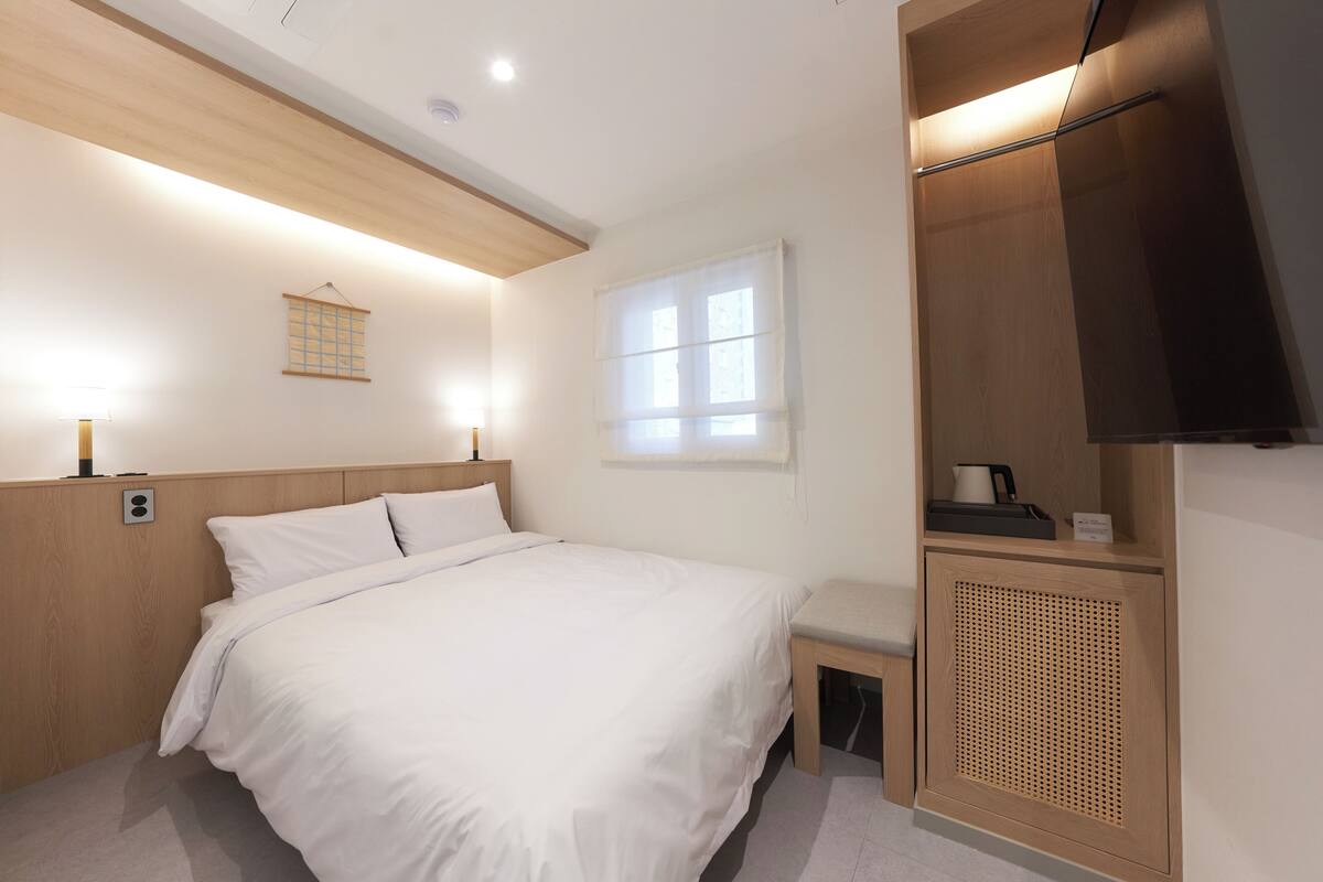 A hotel room with a bed, bedside table, a small bench, a window with blinds, a wall-mounted TV, and a wooden cabinet.