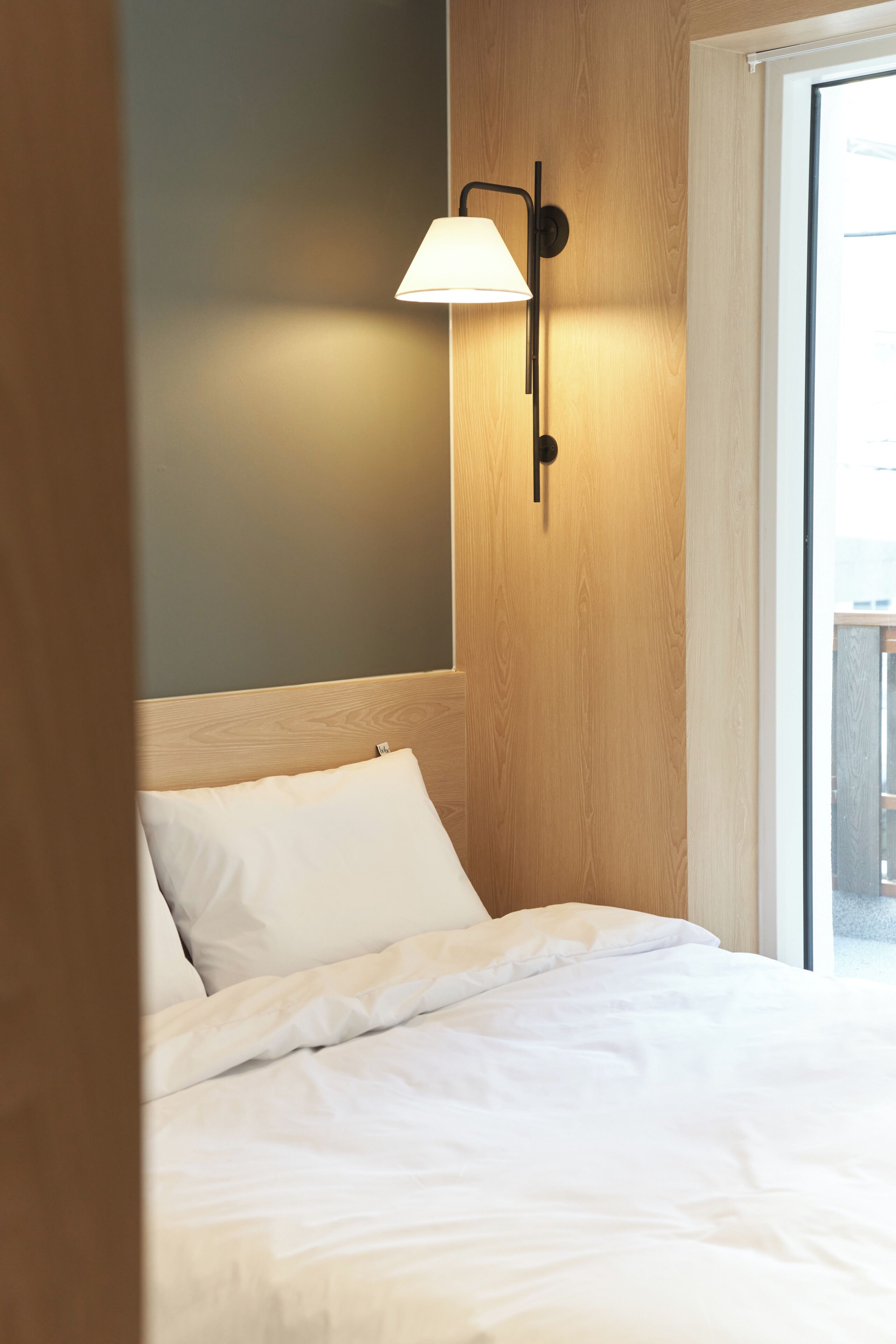 A hotel room with a bed, a wall-mounted lamp, and a window.