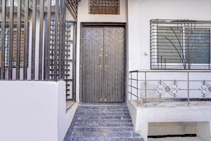 A staircase leading to a door with a metal gate on the left, a window with a metal grille on the right, and a white wall. - Classic Hotel Ramashray Palace (Ujjain)