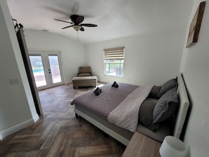 A bedroom with a bed, a chair, a ceiling fan, a window with blinds, and a view of a pool. - IMG Retreat • Heated Pool • 4BR (Bradenton)