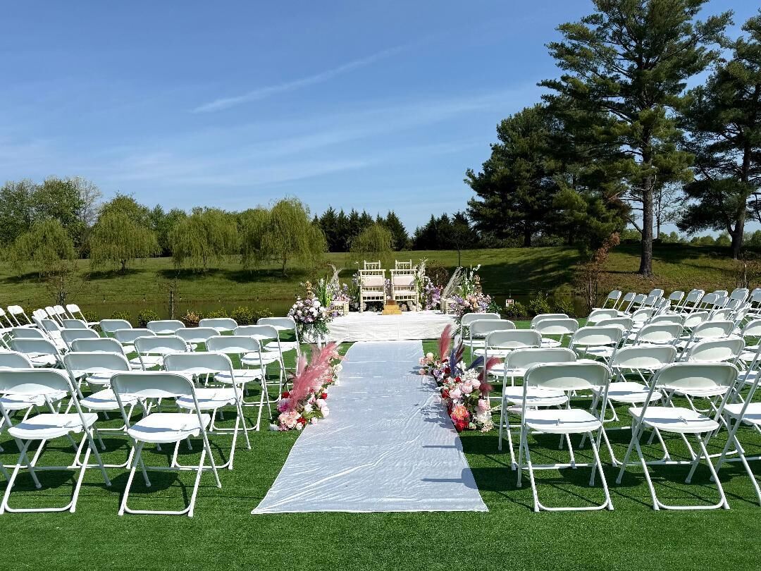 An outdoor wedding ceremony setup with white chairs arranged in rows, a white aisle, and floral decorations.