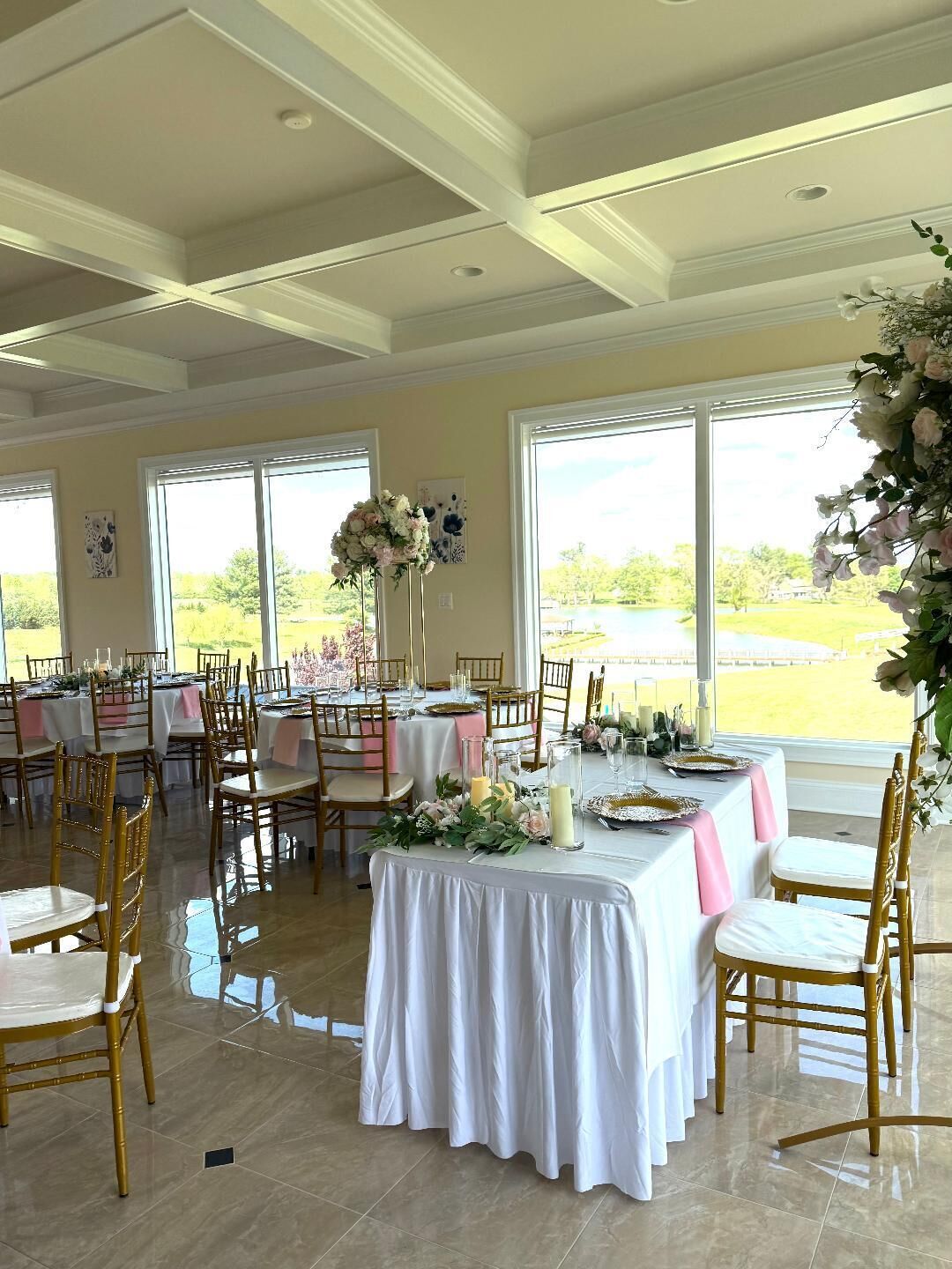 Une salle de banquet avec des tables dressées pour un événement formel, de grandes fenêtres et une vue sur l’extérieur.