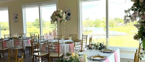 Une salle de banquet avec des tables dressées pour un événement formel, de grandes fenêtres et une vue sur l’extérieur.