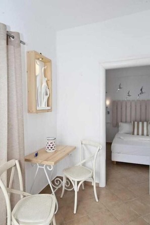 Una habitación con una cama, un escritorio con dos sillas, una mesita con un objeto decorativo y una obra de arte colgada en la pared.
