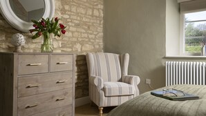 Ein Schlafzimmer mit einem hölzernen Schminktisch, einem gestreiften Sessel, einem Bett mit grüner Bettdecke, einem Fenster mit Aussicht und einer Heizung.