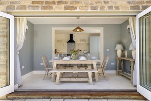 A dining area with a wooden table set for four, a hanging light fixture, and a kitchen in the background. - Primrose Cottage, Barnsley -  beautiful dog friendly cottage with hot tub (Barnsley)
