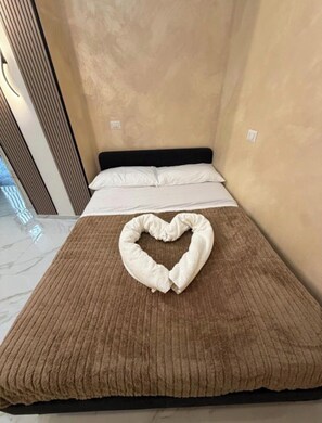A bed with a heart-shaped towel on the bedspread, a black headboard, and a beige wall. - HouseNaplesCenter (Napoli)