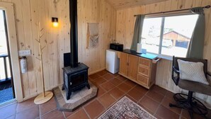 Een houten interieur met een open haard, een kleine koelkast, een raam met gordijnen en een stoel.