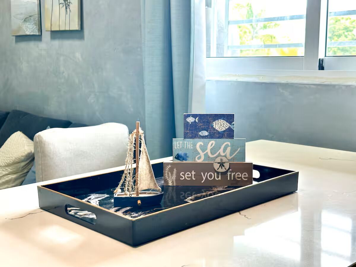 Una bandeja con un modelo de barco y un letrero decorativo sobre una mesa en una habitación con ventana y sofá.