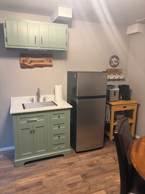 Une cuisine avec des meubles de couleur verte, un réfrigérateur en acier inoxydable, un panneau indiquant l’emplacement du coin café et une table en bois.