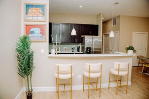 Cocina moderna con gabinetes oscuros, un refrigerador de acero inoxidable y una encimera blanca. Hay tres taburetes blancos y una planta en maceta.