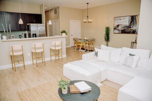 Una sala de estar moderna con un sofá blanco, un comedor con mesa y sillas, una cocina con armarios oscuros y un refrigerador, y un cuadro en la pared.