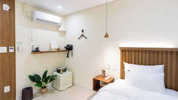 A hotel room with a bed, bedside table, hanging light, air conditioning unit, and a small desk with a plant.