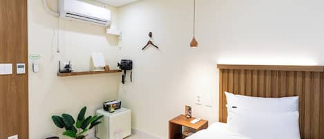 A hotel room with a bed, bedside table, hanging light, air conditioning unit, and a small desk with a plant.