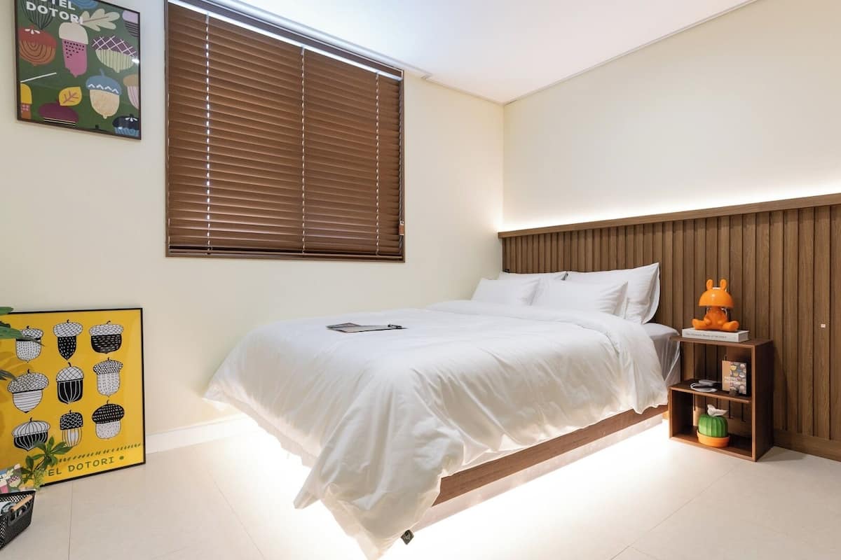 Un dormitorio con una cama, una mesita de noche, un estante en la pared y una ventana con persianas.