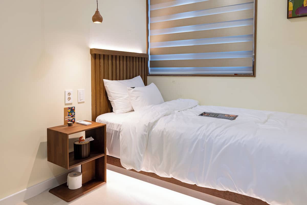Un dormitorio con una cama, una mesita de noche, una lámpara, una ventana con persianas y un estante en la pared.