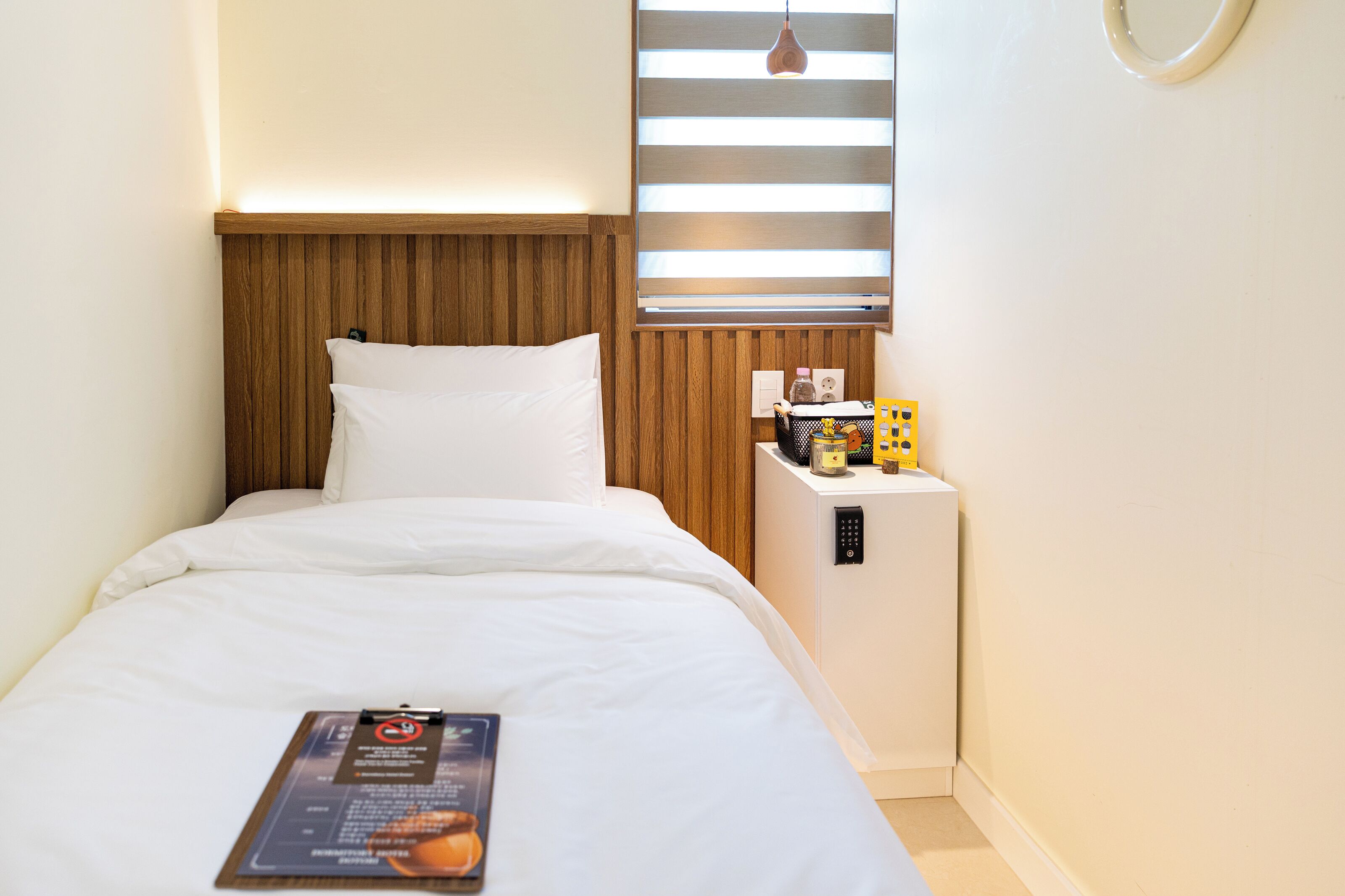A hotel room with a bed, a nightstand with a lamp, a small table, and a window with blinds.