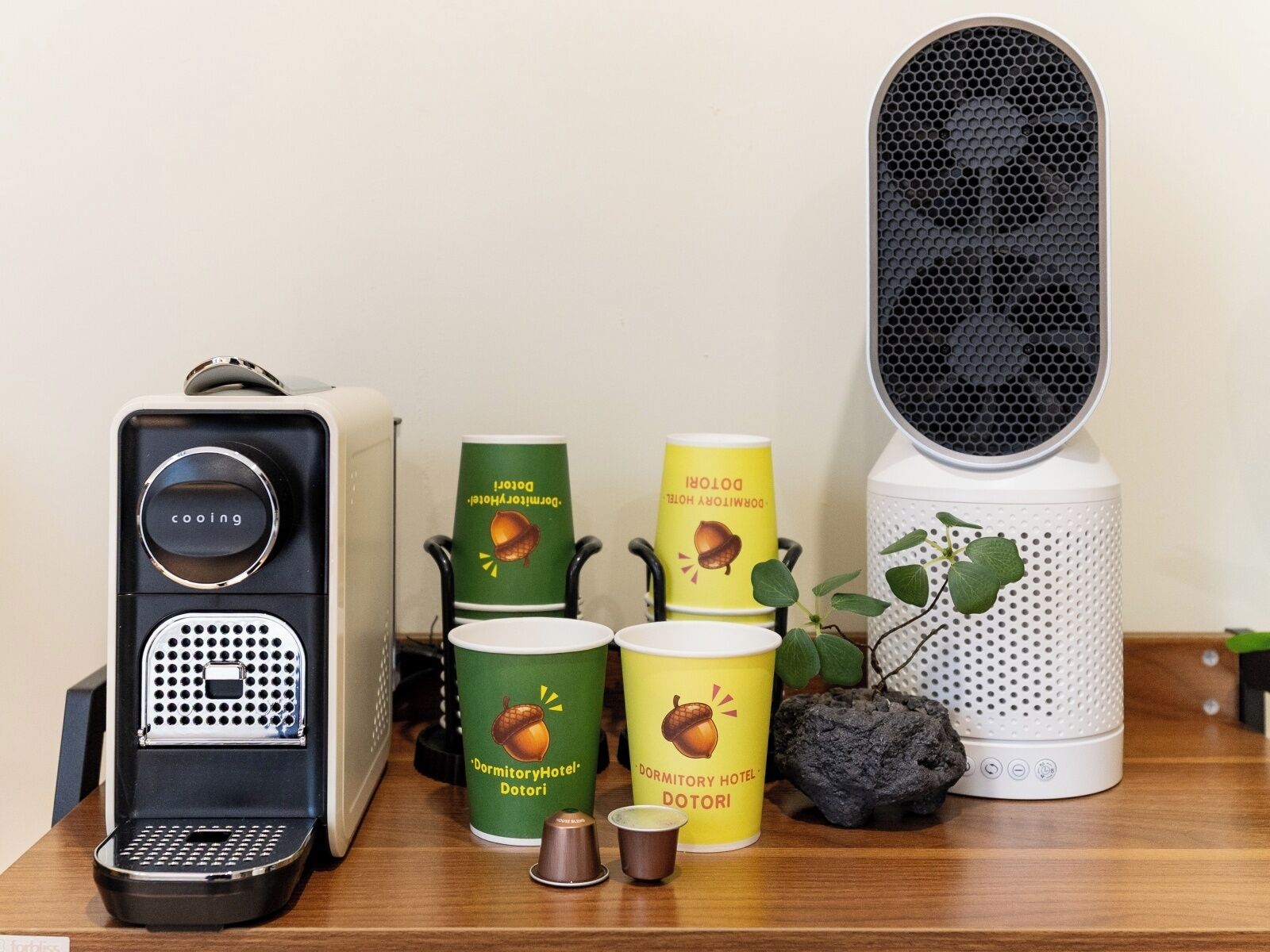 A coffee machine, two cups with 'Dormitory Hotel Dotori' branding, a speaker, and a small plant on a wooden surface.