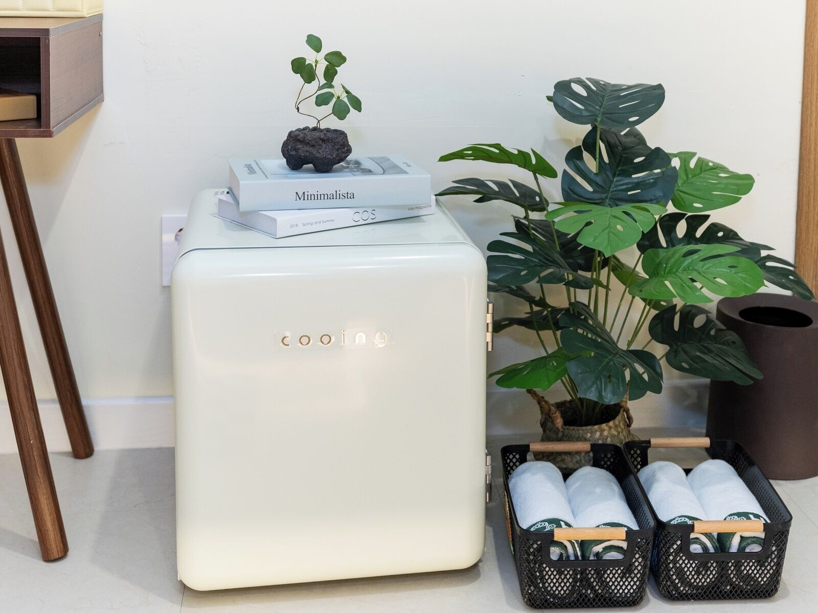 A white mini-fridge with 'cooling' branding, a small potted plant, a magazine, and a black basket with towels.