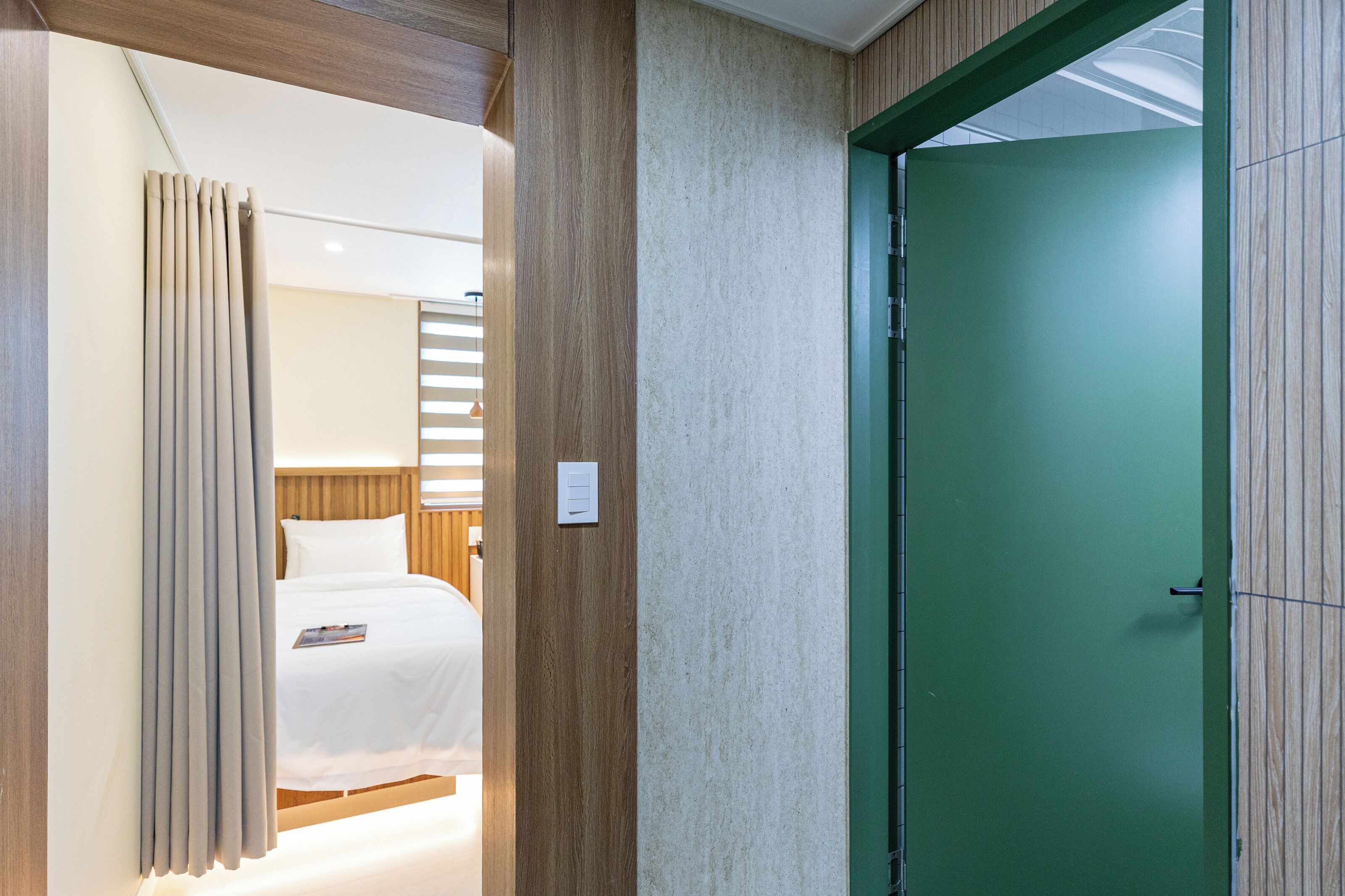 A hotel room with a bed, curtains, a green door, and a light switch on the wall.