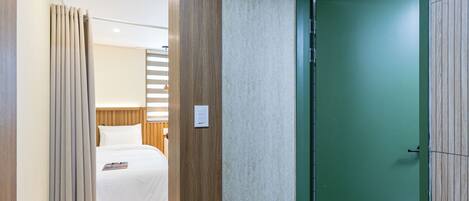 A hotel room with a bed, curtains, a green door, and a light switch on the wall.