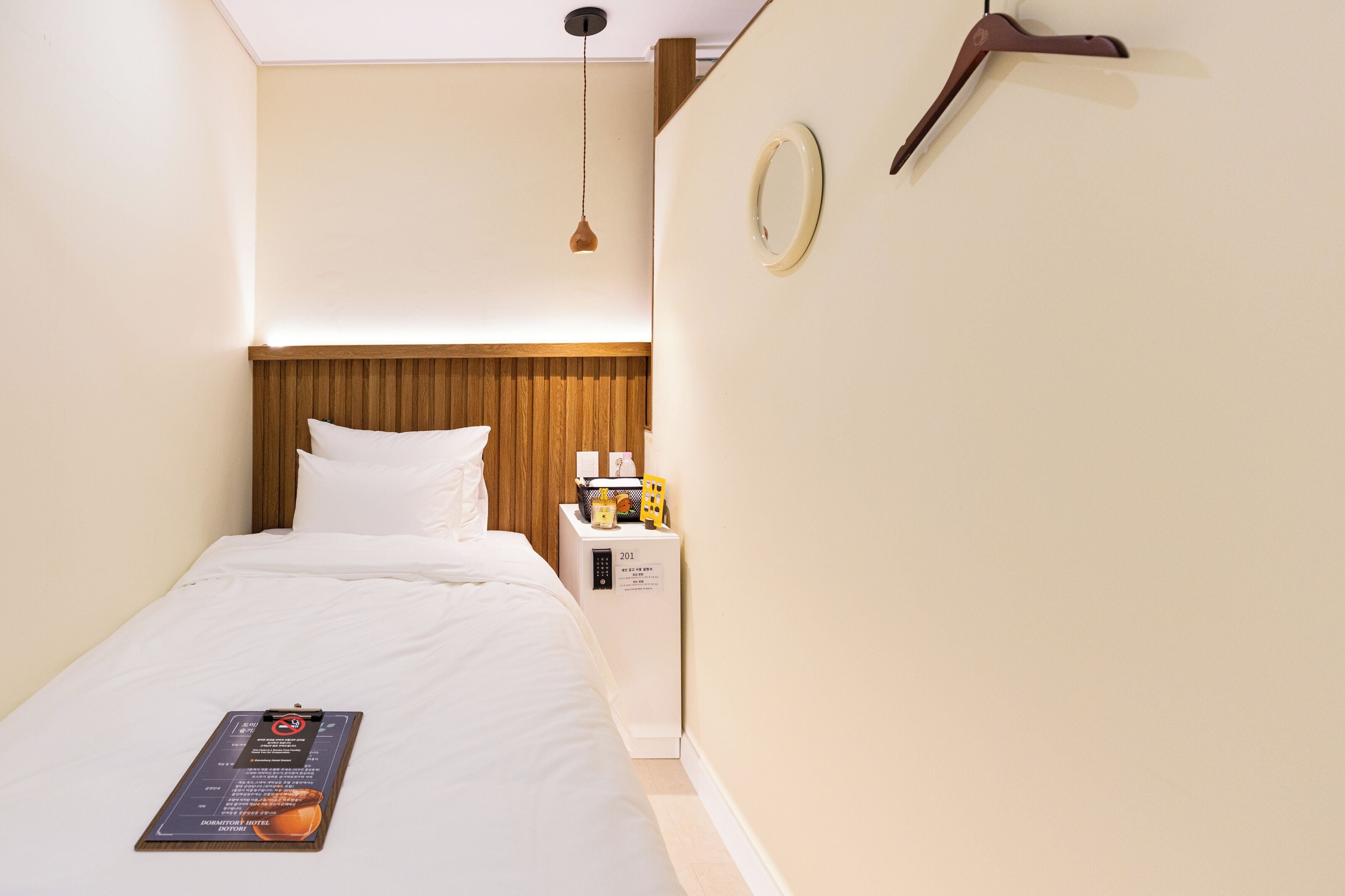 A hotel room with a bed, a bedside table with a lamp, a wall-mounted mirror, and a door with a number plate.