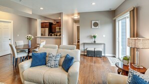 A modern living room with a sofa, coffee table, and a dining area with chairs. There is a kitchen in the background with cabinets and appliances. - 4 Mi to Easter Lake Park! Inviting Rental w/ Patio (Des Moines)