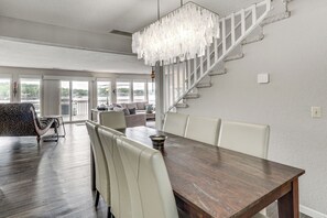 Un espace repas comprenant une table en bois, des chaises blanches, un lustre et un escalier avec une rampe blanche.
