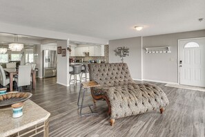 Un salon avec un fauteuil à capitons, un coin repas avec une table et des chaises, et une cuisine en arrière-plan.