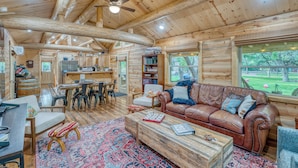 A log cabin interior with a dining area, a leather sofa, and a large window overlooking a yard. - Blanco Riverfront Estate w/ 2 Homes: 4-Acre Oasis (Blanco)