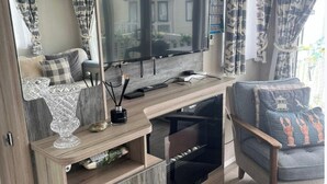 Un área de estar con televisor, una mesa de centro de madera, una silla con cojín estampado y cortinas de temática náutica.