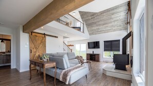 Un vaste espace de vie comprenant un coin nuit en mezzanine, un plancher en bois, un canapé gris et une cheminée.