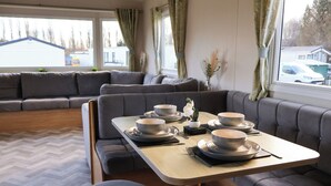 A modern living room with a dining area, grey sofas, a wooden table set with bowls and plates, and large windows with curtains.