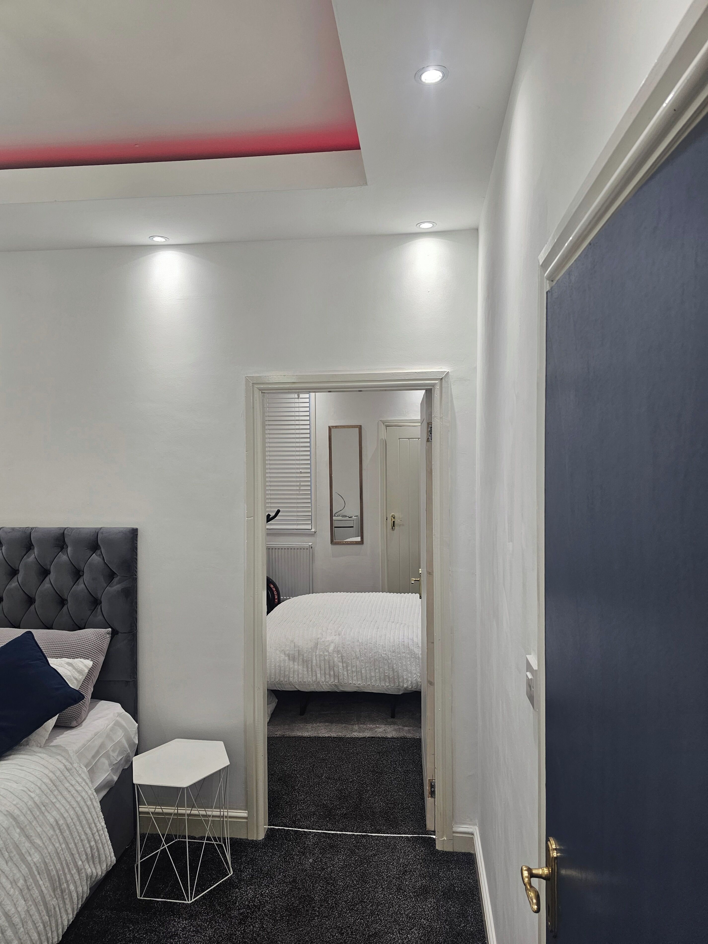A bedroom with a bed, a small side table, a door, and a hallway leading to another room.