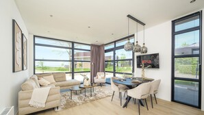 Un moderno salón con sofá, mesa de comedor y sillas, ventanas amplias y vistas al exterior.