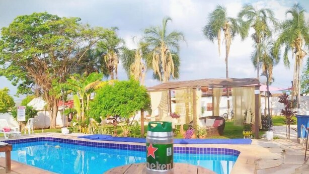 A pool area with a wooden table and stools, surrounded by palm trees and a building with a covered area. - Chalés Bela Vista (Pirenopolis)
