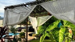 A covered outdoor seating area with a table, a potted plant, and a view of a pool and greenery. - Chalés Bela Vista (Pirenopolis)