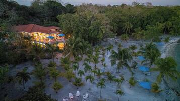 A beachfront property with a large house, a fire pit surrounded by lounge chairs, and a sandy beach with palm trees.