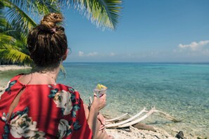 Una persona sosté una beguda a la vora d'una platja amb aigua cristal·lina i una palmera.