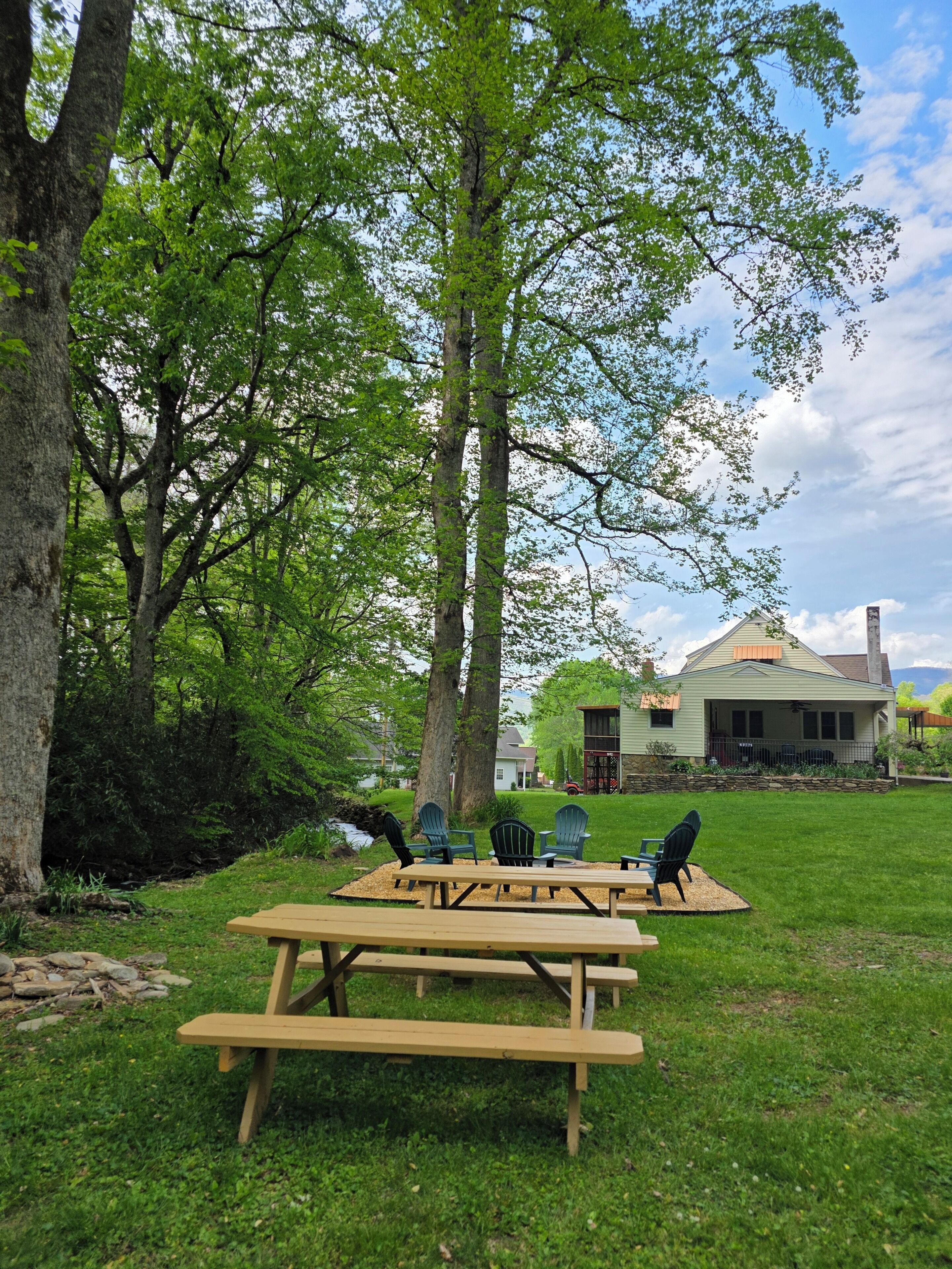 A picnic area with benches and chairs, surrounded by trees and a house in the background.