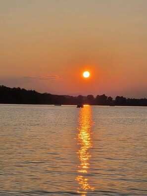 Ein Sonnenuntergang über ruhigem Gewässer, im Vordergrund eine Baumsilhouette und weiter entfernt ein Boot.