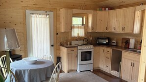 Interior de una cabaña de madera con una mesa de comedor, sillas, una zona de cocina y un ventilador de techo.