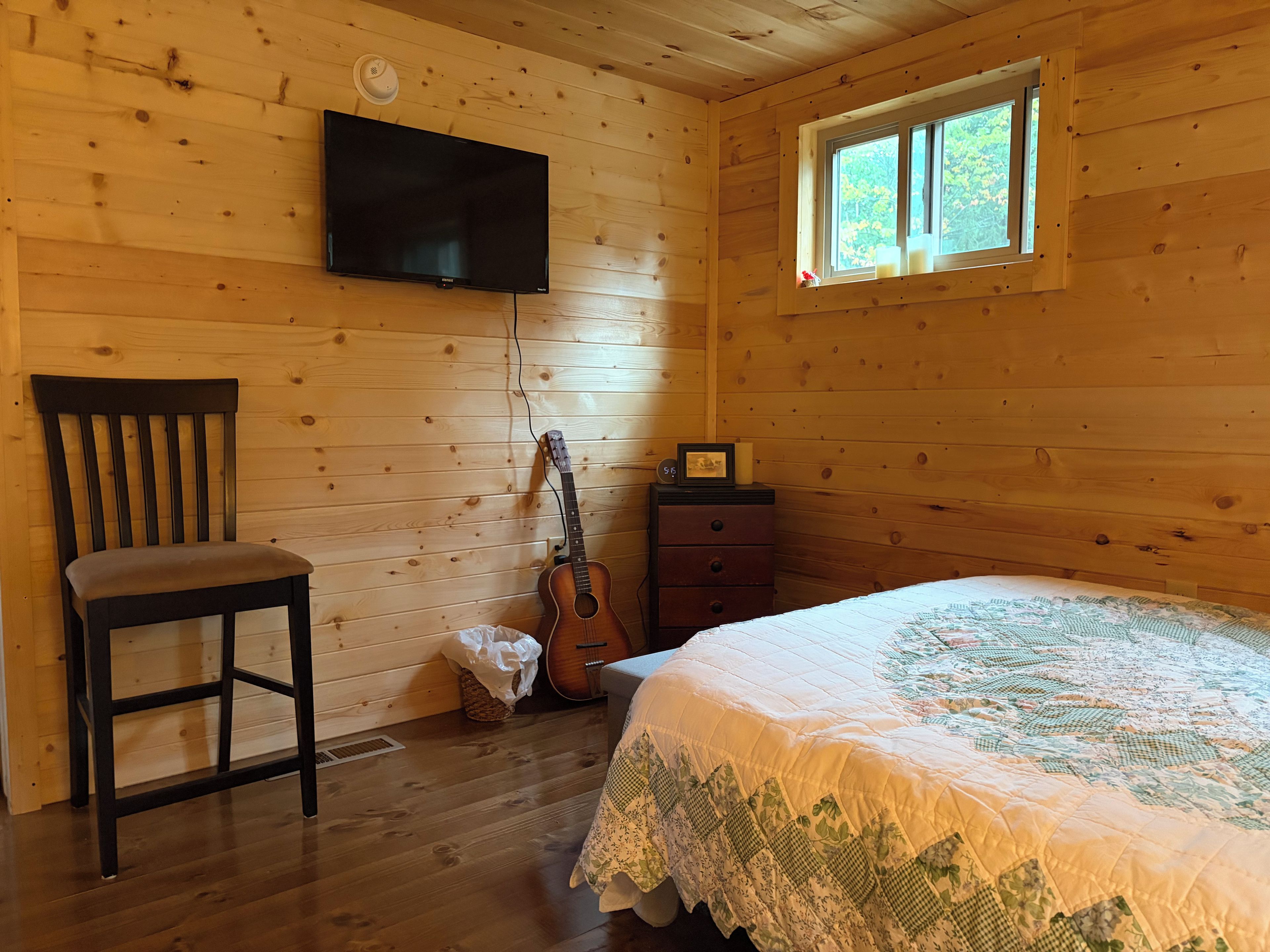 Une pièce de style cabane en bois, comprenant un lit, une chaise, une guitare, une commode et un téléviseur fixé au mur.