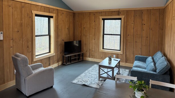 Une pièce aux murs recouverts de lambris en bois, un téléviseur posé sur un meuble, un fauteuil gris, un canapé bleu, une petite table et une fenêtre.