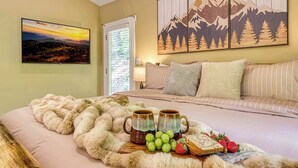 A bedroom with a bed, a painting of mountains, a window with blinds, and a tray with fruit and mugs. - Pine and Peak – Hot Tub, Fire Pit, Dog Friendly (Gatlinburg)